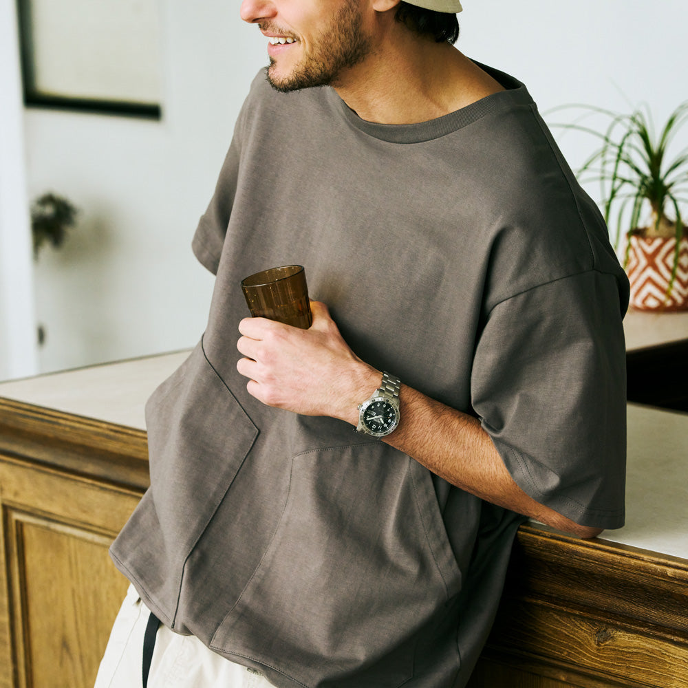 Man wearing a gray oversized shirt holding a wooden cup indoors.