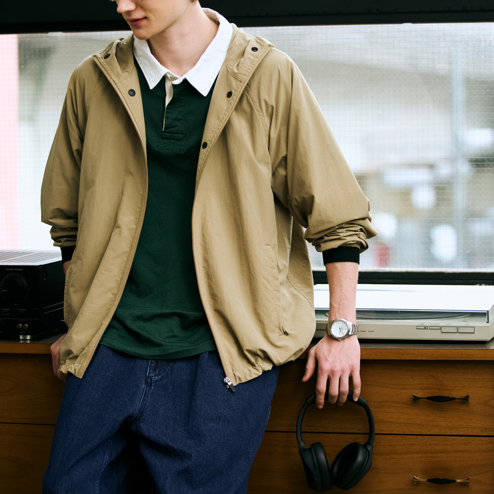 Person wearing a beige jacket over a green shirt with headphones on a wooden surface.