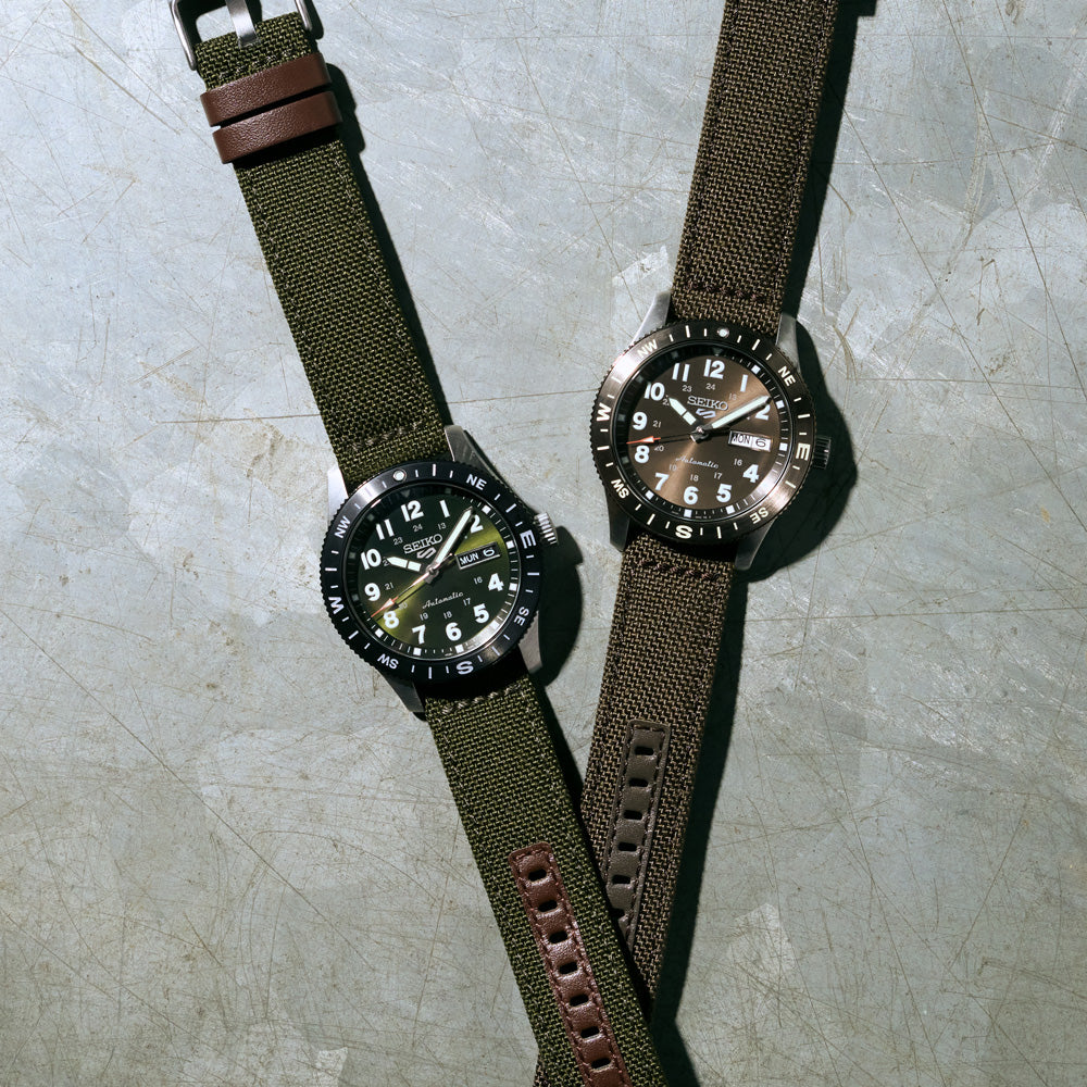 Two watches with green and brown straps on a textured surface
