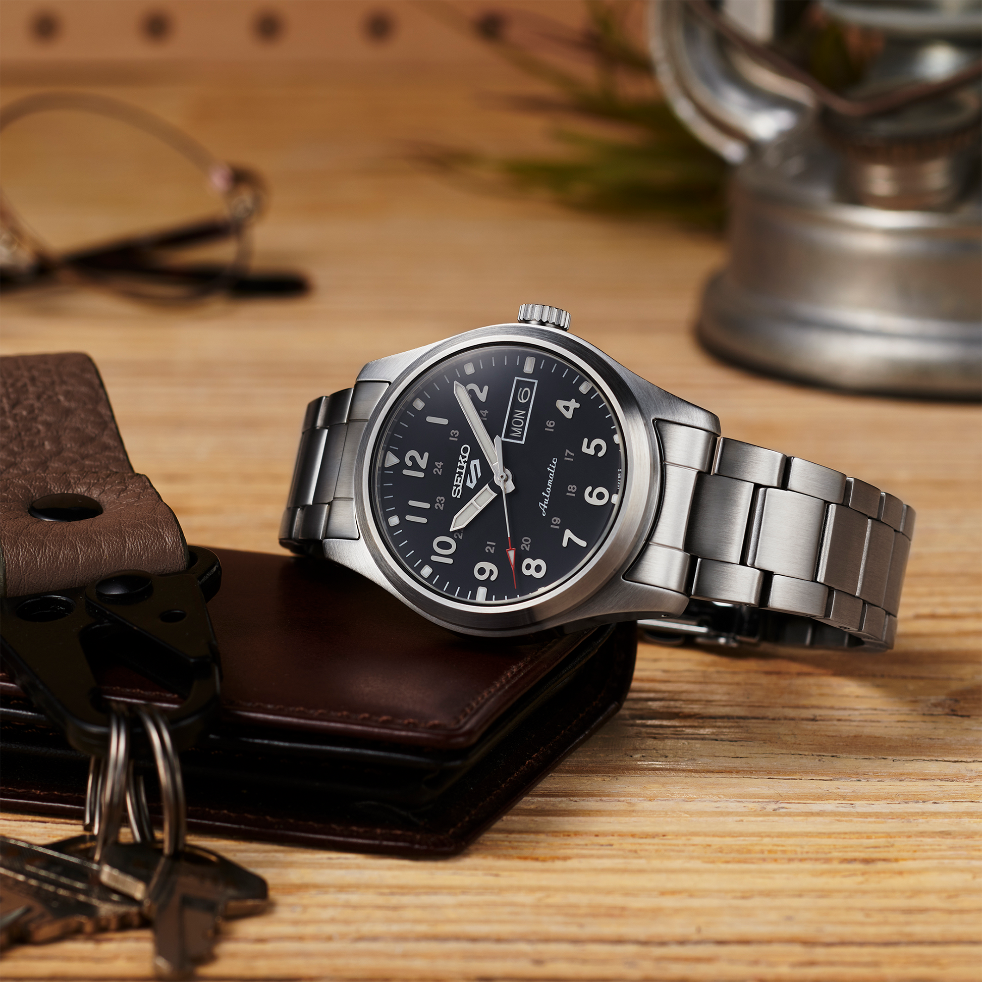 Silver wristwatch with black dial on a wooden surface with wallet and keys.