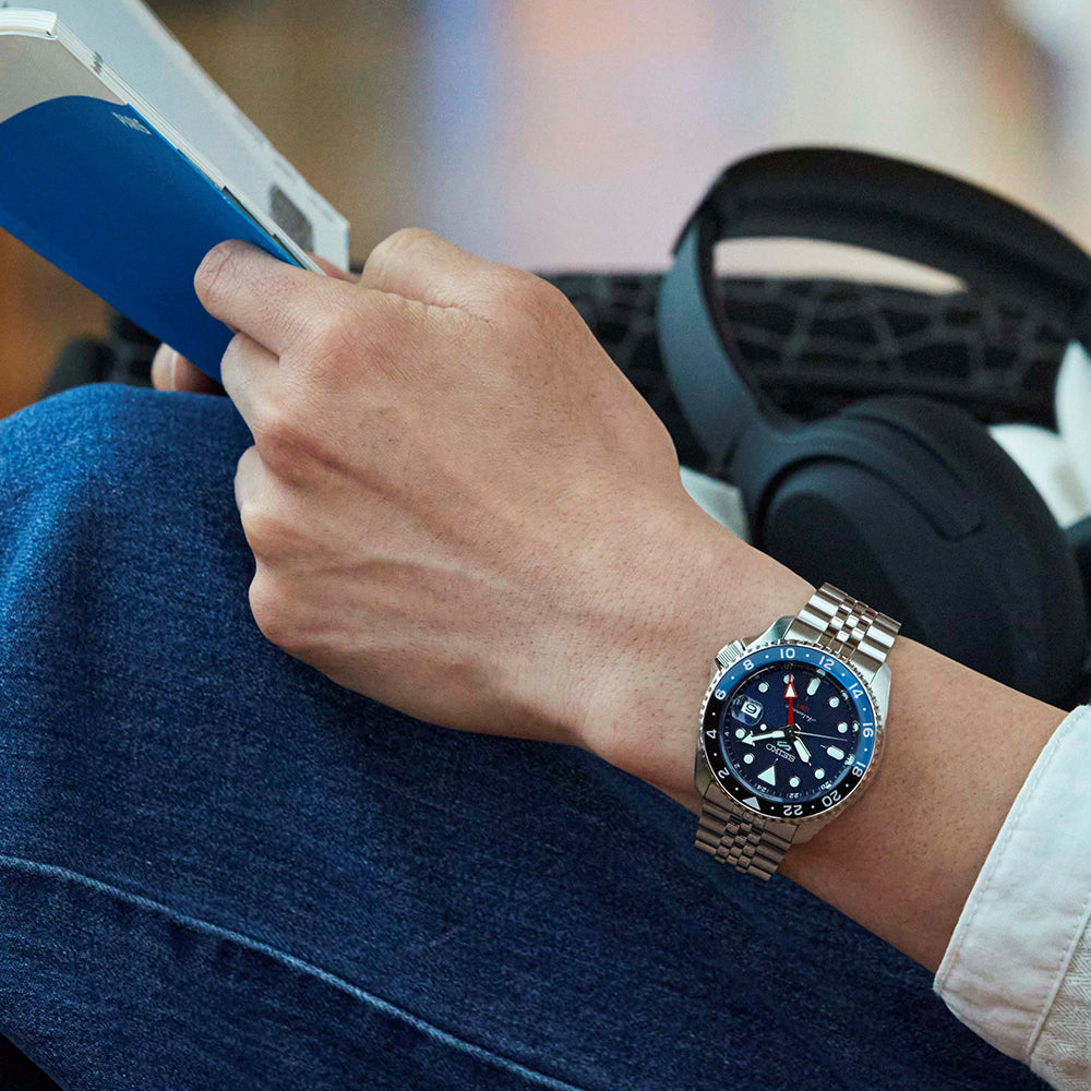 Hand wearing a watch with a blurred background of a person using a smartphone.