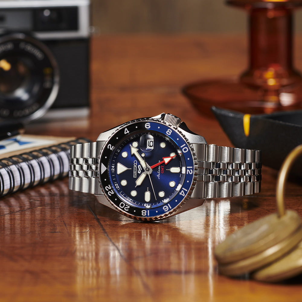 Silver watch with a blue face on a wooden surface with a camera and other items in the background