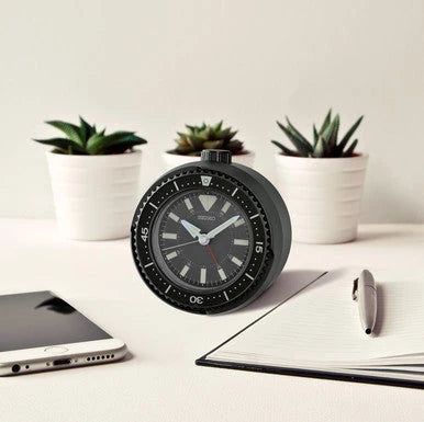 SEIKO QHE207KLH rotating bezel desk alarm clock displayed on a workspace with small plants, notebook, pen, and smartphone.