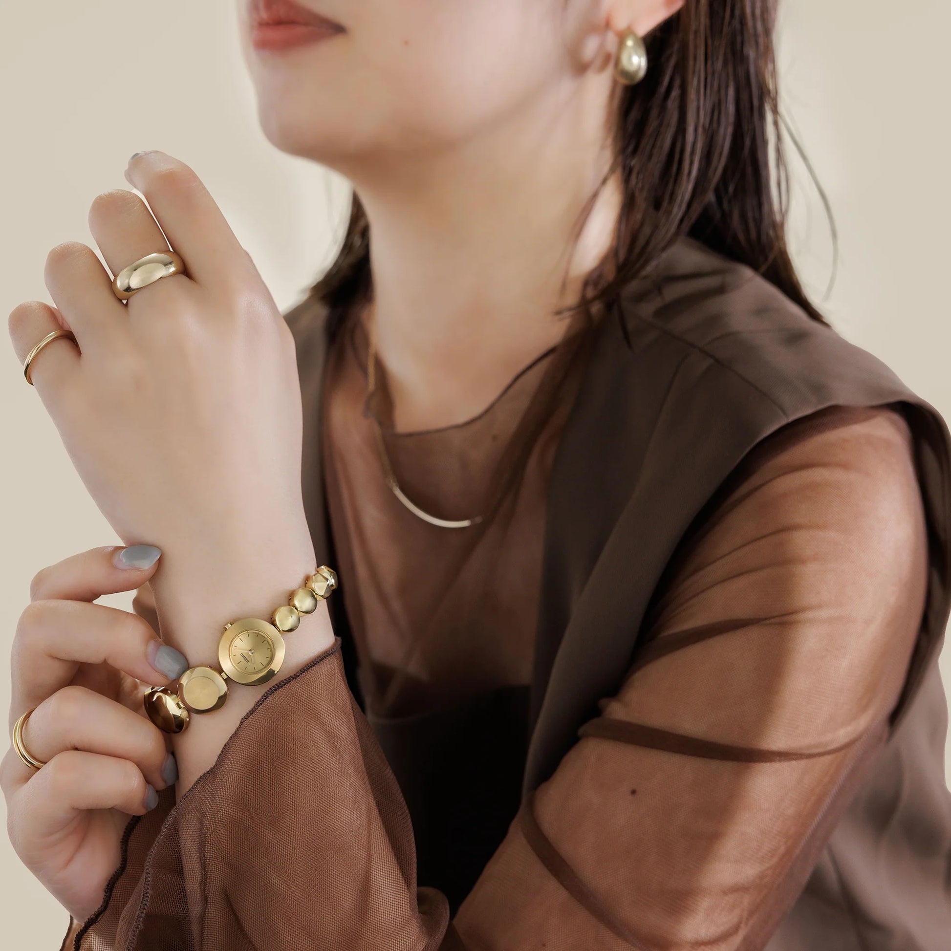 Close-up of a person wearing gold jewelry including rings, a bracelet, and earrings on a neutral background.