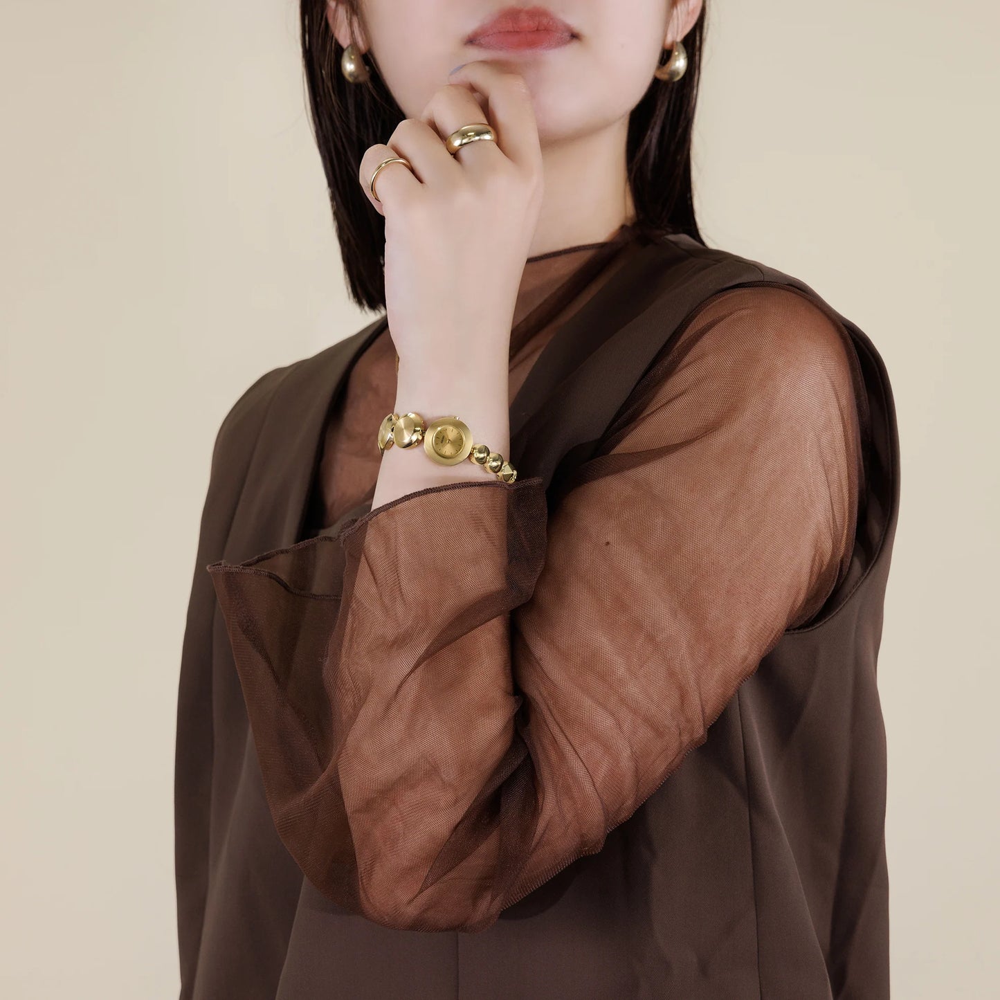Woman wearing a brown sheer top with gold jewelry on a beige background