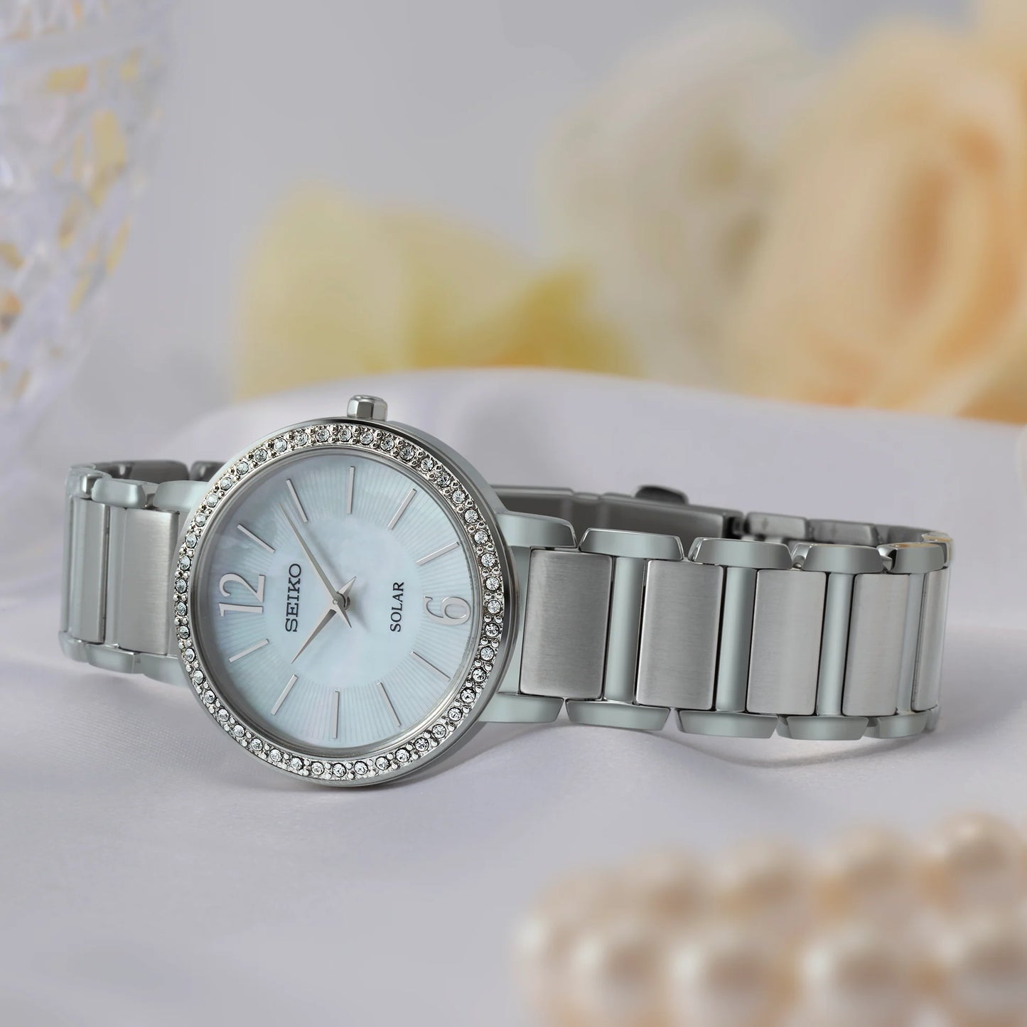 Silver Seiko watch with a decorative band on a white surface with blurred pearls in the background