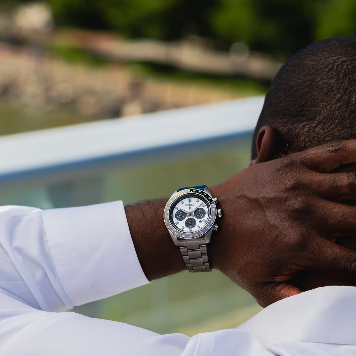A silver stainless steel chronograph watch with a white dial and black bezel, worn on the wrist of a man in a white shirt, photographed outdoors with a blurred background.