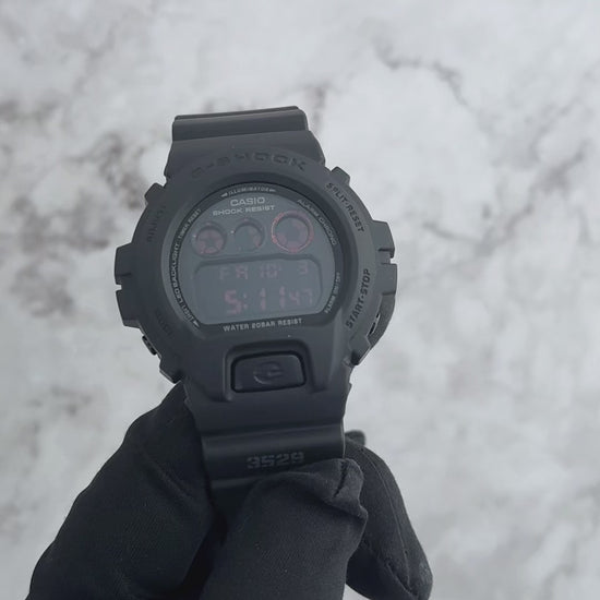 Casio G-SHOCK DW6900MS-1 digital watch in stealth matte black resin case and band with red accents and black ion-plated buttons. Features shock resistance, mineral glass, 200-meter water resistance, electro-luminescent blue-green backlight with afterglow, stopwatch, countdown timer, multi-function alarm, hourly time signal, flash alert, full auto-calendar, and 12/24-hour format. Case size 53.2 × 50 × 18.7 mm, weight 67 g, powered by CR2016 battery with approx. 2-year life.”