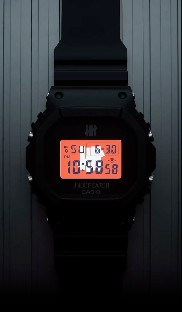 Casio G-SHOCK DWE5600UD-1 UNDEFEATED collaboration model with matte black Carbon Core Guard case, semi-translucent resin band featuring UNDEFEATED logo, green LCD display, engraved caseback, and special packaging. Includes two interchangeable camouflage straps (tiger camo, storm camo). Features shock resistance, mineral glass, 200m water resistance, LED Super Illuminator backlight, stopwatch, countdown timer, alarms, flash alert, auto-calendar, and ±15 sec/month accuracy. Case size 48.9 × 43.8 × 13.7 mm, we